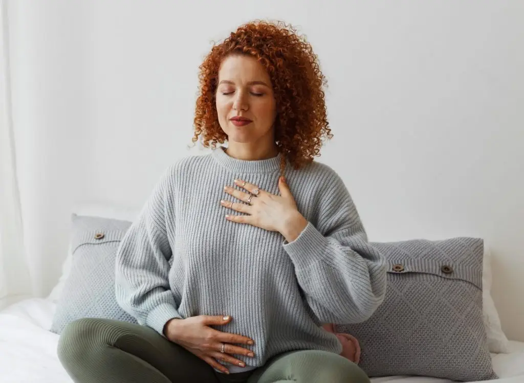 Portrait of plus-size curly redhead female in casual clothes doing pranayama technique sitting on bed next to window with closed eyes, putting hands on chest, breathing slowly and deeply stock photo