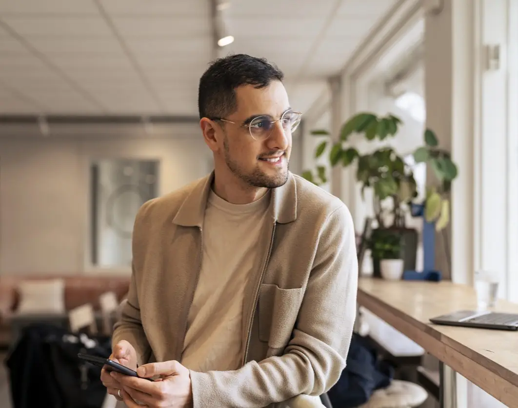 A casually dressed young man wearing glasses sits in a bright cafe workspace, smiling as he checks his smartphone beside an open laptop, conveying relaxed productivity and modern remote work.