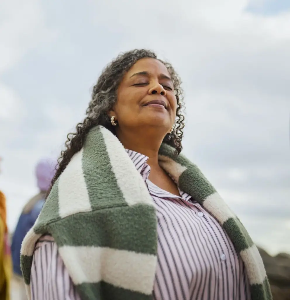 A senior Black woman with curly gray-streaked hair closes her eyes and tilts her face upward in a serene, mindful moment outdoors, wearing a green striped wrap, with friends blurred in the background.