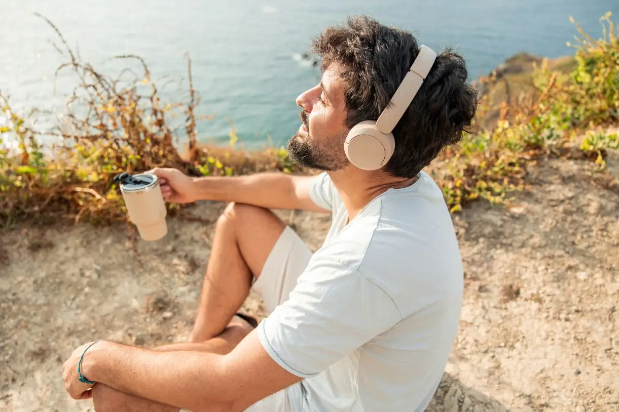 Young adult man sitting on a cliff by the sea, wearing headphones and holding a travel mug, relaxing and enjoying personal time after remote work, dreaming of adventure
